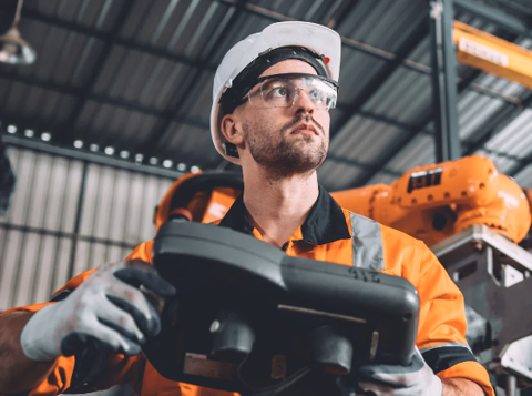 A tradesman working in a warehouse wearing a hi-vis jacket, white gloves, white hard hat and protective safety glasses. He looks out to the distance whilst holding a black control panel in his hands.