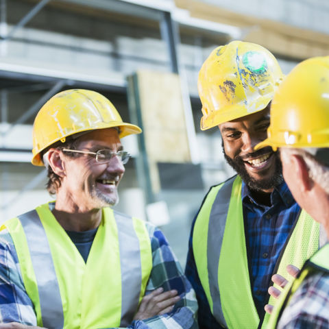 Three construction workers wearing yellow hard hats and high vis vests, standing on a building site, smiling and talking