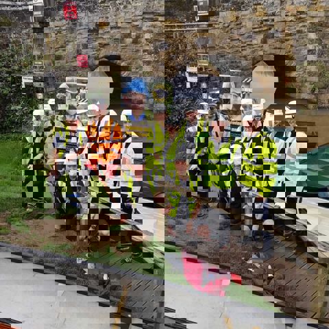 Eleven construction workers wearing hard hats and high vis jackets, standing on a riverbank with a Mates in Mind banner behind them