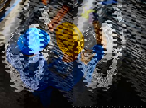 Looking down on two workers wearing hard hats, standing in front of scaffolding, talking about their mental health