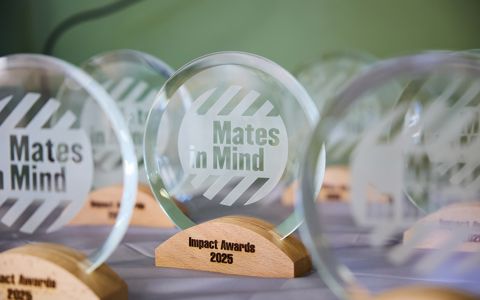 Circular glass trophies with the Mates in Mind logo engraved on it, and standing on a wooden block with Impact Awards 2025.