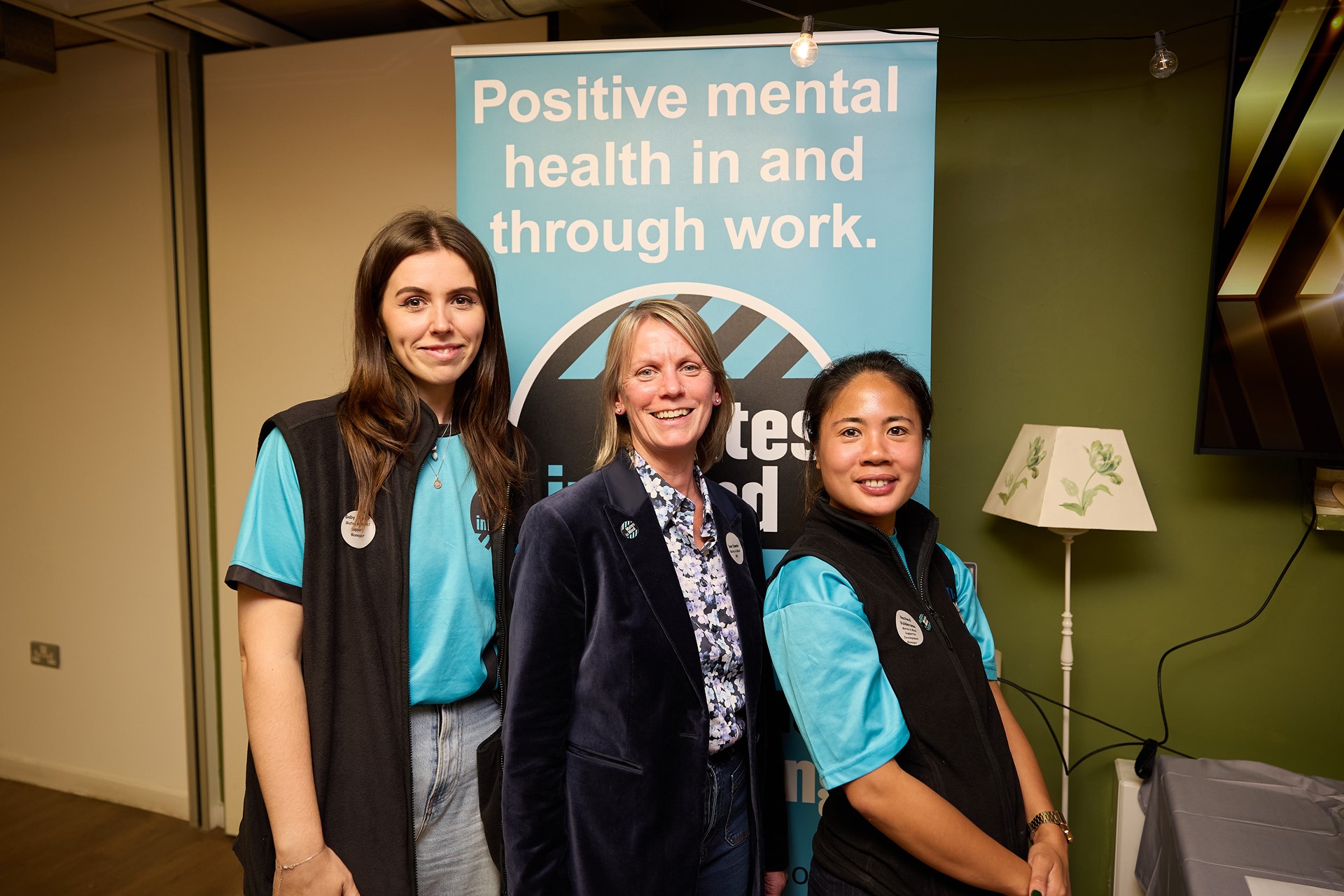 Three members of the Mates in Mind team, standing in front of a banner which says: positive mental health in and through work