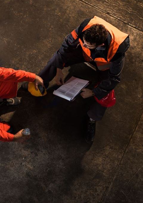 Looking down on two men wearing high vis clothing, sitting on boxes in a warehouse, talking about their mental health.